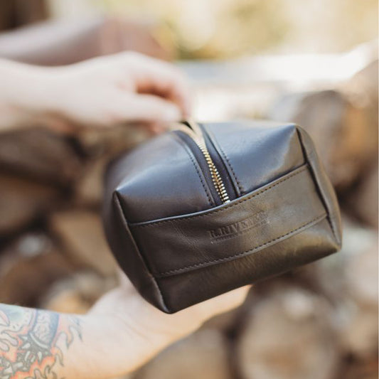 Brown leather toiletry bag with gold zipper held by a person against a blurred natural background