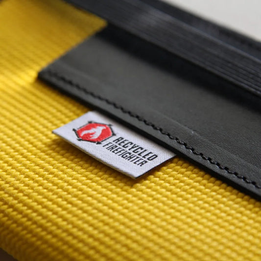 Close-up of The Inspector notebook cover showing decommissioned yellow polyester fire hose texture, top-grain leather pen sleeve, and woven Recycled Firefighter brand label – handmade in USA"