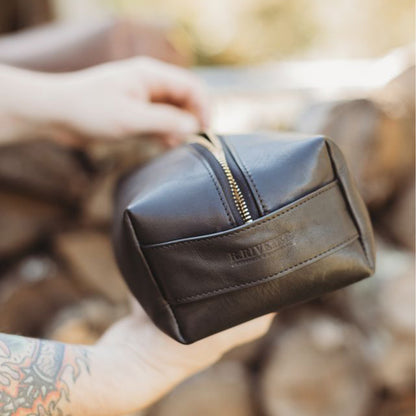 Brown leather toiletry bag with gold zipper held by a person against a blurred natural background