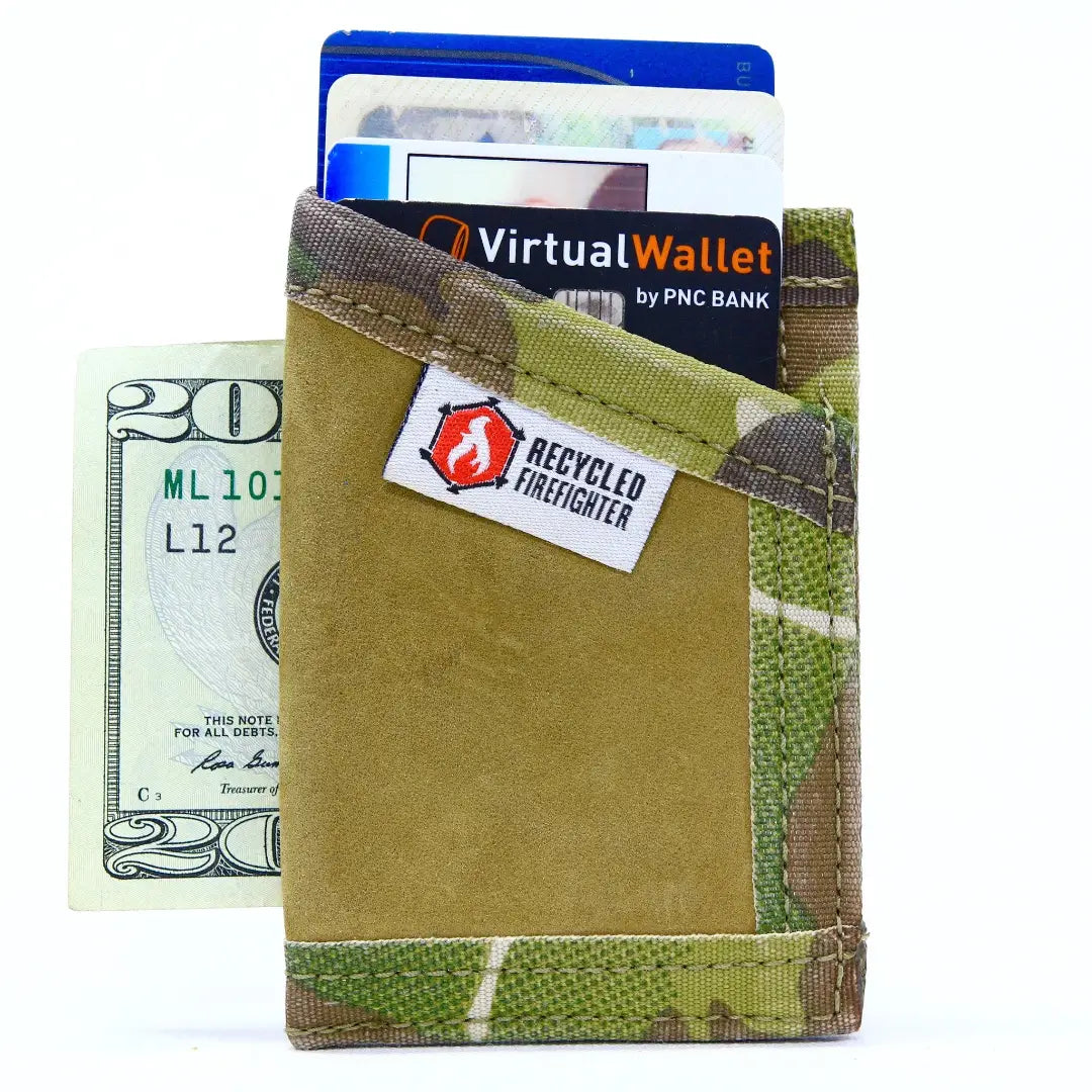 A camouflage-patterned bifold wallet with a tan suede-like center panel, shown holding cards and a dollar bill. A white label on the front reads 'RECYCLED FIREFIGHTER' with a red flame logo.