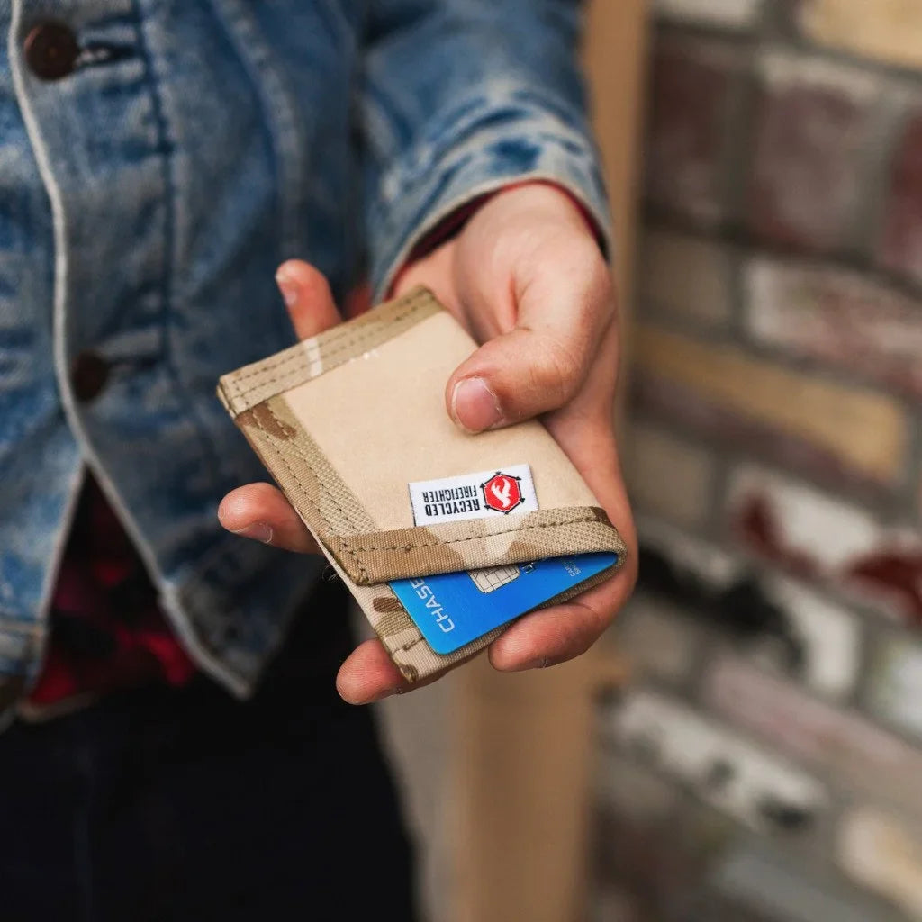 A person in a blue denim jacket holds a beige and brown leather cardholder wallet with a clear ID window and a visible 'RECYCLED' logo patch. A blue credit card is partially visible inside.